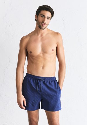 Tânăr cu păr închis la culoare stă fără tricou lângă un perete alb, purtând pantaloni scurți de baie bleumarin cu model și snur.