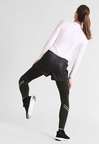 Idrottsensemble bestående av en vit långärmad topp, svarta lädershorts och gröna leggings med reflekterande grå ränder, i kombination med svarta sneakers.