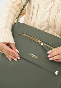 Borsa verde texturizzata con zip oro e logo in evidenza. Presenta una tracolla removibile e una tasca anteriore con zip.
