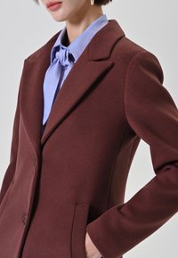 Donna che indossa un cappotto di lana marrone scuro attillato sopra una camicia colletto blu chiaro con un fiocco annodato al collo, in piedi con le mani in tasca.