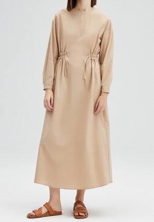 Femme portant une robe midi beige à manches longues avec des détails noués à la taille et des sandales plates couleur tan sur un fond blanc uni.