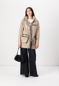 Beżowa kurtka przeciwwiatrowa z kapturem, zasuwaną kieszenią na piersi i podwójnymi kieszeniami z przodu. Czarna torebka oraz szerokie dżinsowe spodnie dopełniają stylizację.