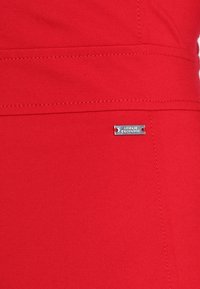Červená tkanina s vodorovným švem a malou obdélníkovou stříbrnou kovovou značkou s vyrytým logem Armani Exchange.