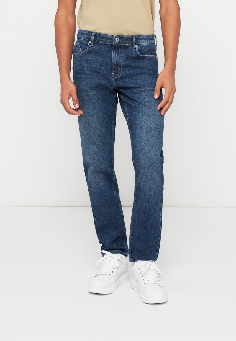 Tamsiai mėlynos džinsinės kelnės su tiesia kojų forma, penkiais kišenėmis ir užtrauktuku. Dėvimos su baltais sportbačiais ir smėlio spalvos marškinėliais su apvaliu kaklu.