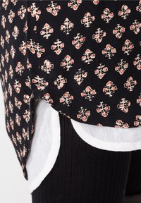 Czarna bluzka z wzorem kwiatowym w kolorze czerwonym i białym, z okrągłym dołem i warstwowym białym wykończeniem na dole, w zestawieniu z czarnymi legginsami w prążki.