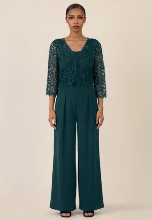 Frau trägt ein dunkelgrünes, kurz geschnittenes Spitzen-Cardigan über einem passenden ärmellosen, weit geschnittenen Jumpsuit, steht vor einem beigen Hintergrund.