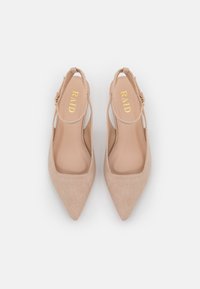 Zapatos de tacón de ante beige con punta afilada y lados abiertos, que presentan un logo dorado en la plantilla y delgadas correas para cerrar en el tobillo.