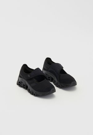 Paar zwarte mesh sportschoenen met dikke, geventileerde zolen en klittenbandsluitingen bovenop, op een effen achtergrond.