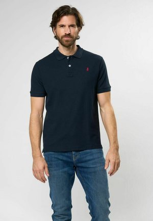 Marineblaues Poloshirt mit klassischem Kragen, Drei-Knopf-Leiste, kurzen Ärmeln und kleinem rotem Logo auf der Brust. Kombiniert mit blauen Jeans.