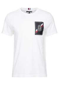 Camiseta de algodón blanca con mangas cortas y un bolsillo en el pecho. El bolsillo tiene un contorno negro con rayas rojas y azules y un diseño de escudo.