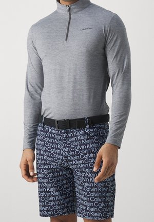 Man wearing grey long-sleeve quarter-zip top and navy shorts with repeating Calvin Klein white logo pattern, black woven belt visible.