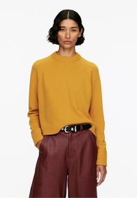 Senfgelbes Strickpullover mit Rundhalsausschnitt, gerippten Bündchen und Saum, kombiniert mit burgunderfarbenen, weiten Lederhosen und einem schwarzen Gürtel.