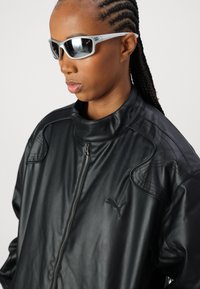 Veste en faux cuir noir avec un col haut, une fermeture éclair et des détails de coutures subtils. Présente un petit logo brodé sur la poitrine.