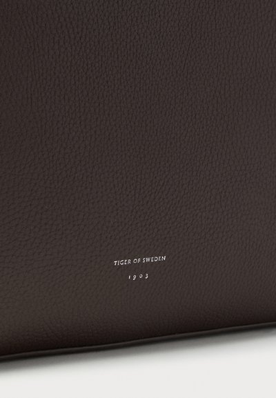 Καφέ σκούρο δερμάτινο πορτοφόλι με υφή στην επιφάνεια, με τον λογότυπο "TIGER OF SWEDEN 1903" ανάγλυφο σε ασημί στο μπροστινό μέρος.