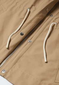 Beige takki, jossa on valkoiset narut, metallinen vetoketju osittain auki, ja hopeiset nepparit vetoketjun molemmin puolin.