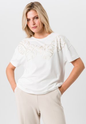 Witte katoenen t-shirt met gouden geborduurde monstera bladeren over de borst, met korte mouwen en een relaxte pasvorm.