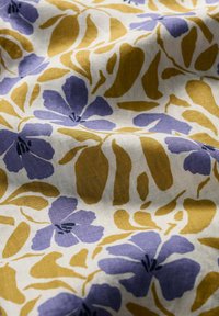 Blommigt tyg med stora, abstrakta violetta blommor och senapsgula blad på en krämfärgad bakgrund; mjuk textur, lätt material.