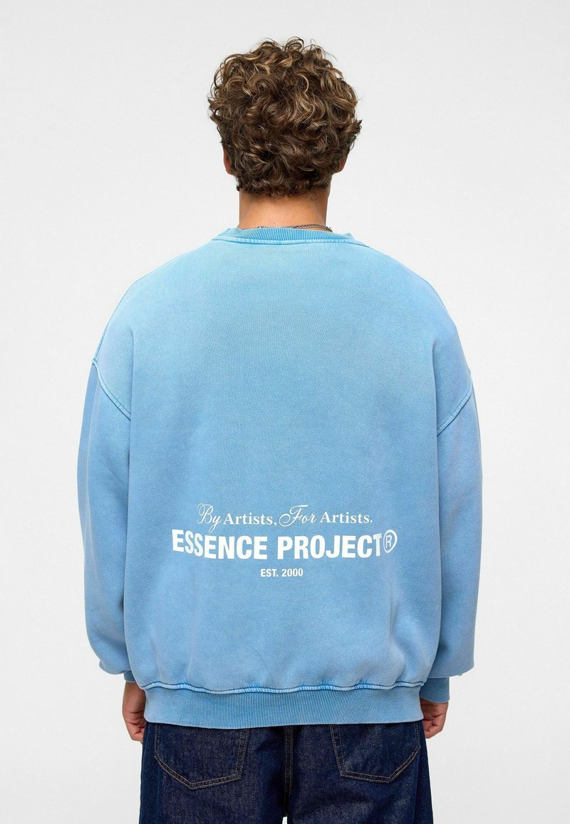 Blauwe sweatshirt met een ontspannen pasvorm, gemaakt van zachte stof. Opvallende witte tekst op de achterkant: "ESSENCE PROJECT EST. 2000."