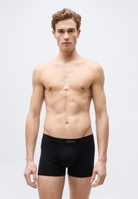 Νεαρός άντρας με σγουρά μαλλιά που φοράει μαύρα μπόξερ DIM, στέκεται μπροστά σε απλό ανοιχτό φόντο, κοιτάζοντας μπροστά με ουδέτερη έκφραση.