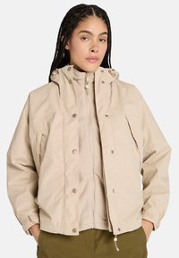Chaqueta impermeable beige con capucha, que cuenta con botones a presión, una cremallera frontal y una capa interior de forro polar. Textura suave y corte relajado.