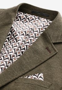 Blazer marrón tejido con forro blanco y marrón de patrón. Presenta solapas de muesca, un bolsillo para el ticket y una textura suave.