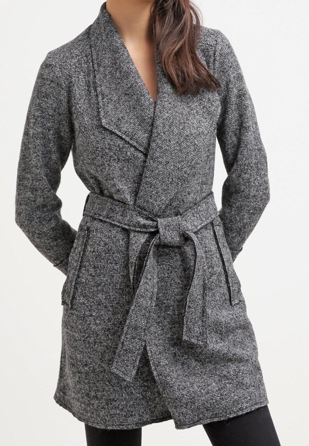 Femme portant un manteau en laine gris texturé avec ceinture, larges revers et poches latérales, mains placées derrière le dos.
