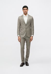 Abito a quadri marrone con giacca monopetto, rever a lancia e pantaloni abbinati. Abbinato a una camicia bianca e scarpe di pelle scura.