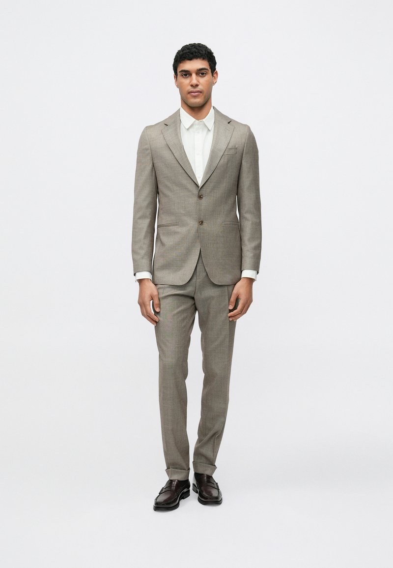 Abito a quadri marrone con giacca monopetto, rever a lancia e pantaloni abbinati. Abbinato a una camicia bianca e scarpe di pelle scura.