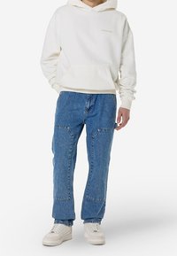 Vaaleanbeige huppari etutaskulla ja pienellä logolla, yhdistettynä sinisiin farkkuihin, joissa on kaksi etutaskua ja rento istuvuus. Valkoiset lenkkarit.