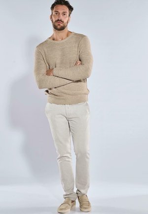 Beige Strickpullover mit weitem Ausschnitt, kombiniert mit hellbeigen Slim-Fit-Hosen und lässigen Slippern, vor einem neutralen Hintergrund stehend.