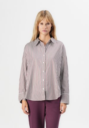 Woman wearing a loose, long-sleeve button-up shirt with brown and white vertical stripes and solid purple pants against a white background.