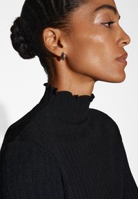 Un col roulé noir à côtes avec une finition texturée et un col volanté. Le mannequin porte une boucle d'oreille ovale et brillante. Fond blanc simple.