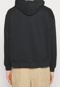 Persona che indossa un hoodie nero Converse con il cappuccio alzato e pantaloni beige Carhartt, mostrata di spalle su uno sfondo neutro.