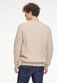 Beige Kabelstrickpullover mit Rundhalsausschnitt und gerippten Bündchen, getragen über dunkelblauen Jeans. Verfügt über ein strukturiertes Muster über die gesamte Fläche.