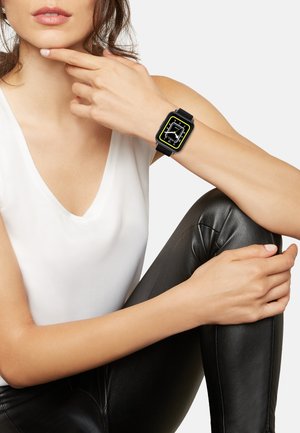 Sort smartwatch med firkantet urskive, gule accenter og hvide markeringer. Båret på et håndled, parret med en hvid top og sorte læderleggings.