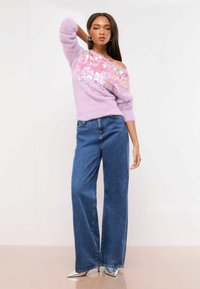 Hellblaues, flauschiges Sweatshirt mit Paillettenverzierung über einer Schulter, kombiniert mit hochgeschnittenen, weiten blauen Denim-Jeans und silbernen High Heels.