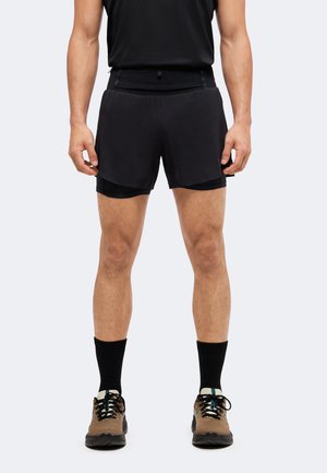 Homme portant un short de course noir superposé, des chaussettes noires, des chaussures de sport marron et noires, debout avec les mains détendues le long du corps.