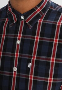 Camicetta a quadri in tonalità navy, rosso e grigio. Presenta un colletto classico, bottoni argentati e una tasca sul petto con un'etichetta del marchio.