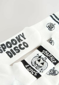 Weiße Socken mit schwarzem Text, der "SPOOKY DISCO" und "SPARKLE & SPICE" zeigt. Die Designs umfassen Kürbisse und ein strukturiertes, geripptes Muster.