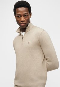 Νεαρός Μαύρος άντρας που φοράει μπεζ πουλόβερ με φερμουάρ στο λαιμό, στέκεται με τα χέρια πίσω από την πλάτη, κοιτάζοντας με αυτοπεποίθηση την κάμερα σε λευκό φόντο.