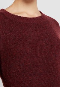 Bordeaux sweater med en ribbet, bred halsudskæring og et blødt, tekstureret strikstof, der fremhæver en hyggelig, afslappet pasform.