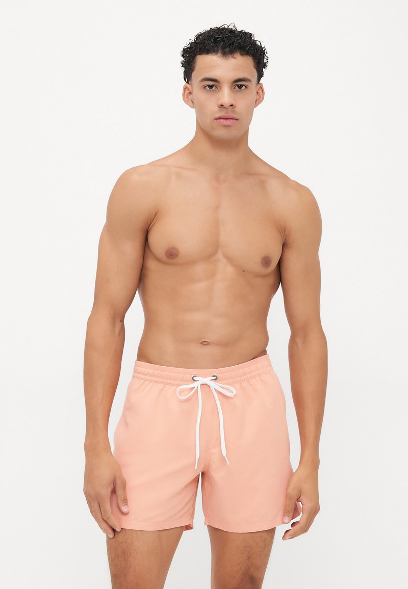 Shorts de bain couleur pêche avec une taille à cordon de serrage, fabriqués en tissu léger, présentant une texture lisse et une coupe classique.