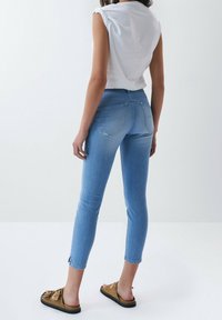 Jeans skinny de mezclilla azul claro con una cintura de media altura, que cuentan con bolsillos traseros y cremalleras en los tobillos. Combinados con una blusa blanca de manga corta y sandalias marrones.