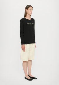 Black long-sleeve shirt with gold embroidered text, paired with cream-colored knee-length shorts and black ballet flats on a plain background.