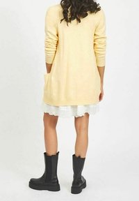 Long yellow cardigan made of soft material with ribbed cuffs and a pocket, worn over a white lace-hem dress and black ankle boots.