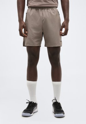 Muž v taupe športových šortkách, bielych ponožkách s lemom a čiernych bežeckých topánkach stojaci na bielom pozadí.