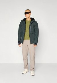Giacca con cappuccio verde scuro con tasche con cerniera, abbinata a una maglietta verde oliva a maniche corte e pantaloni beige chiaro, indossata con occhiali da sole e scarpe da ginnastica.