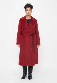 Casaco longo vermelho, com cinto, feito de um tecido macio, apresentando um colarinho largo, dois bolsos frontais e mangas longas. Usado sobre uma blusa preta e calças.