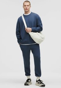 Tengerészkék pamut melegítő szett, amely egy kereknyakú pulcsit és szűkített jogger nadrágot tartalmaz. Krém színű crossbody táskával és fekete sportcipőkkel van párosítva.