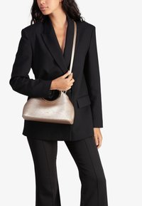 Bolso de mano metálico dorado con un asa redondeada; textura suave; diseño compacto, sostenido por una persona que lleva puesta una chaqueta y pantalones negros.